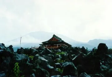 A wooden building sits amid rocky terrain, with a mountain visible in the background under a cloudy sky.