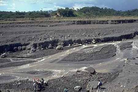Dried up deposit winding through a grey and rocky landscape with a tiny group of people in the foreground contrasting with the size of the former river