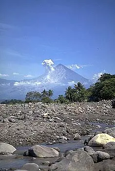A stream flows through a rocky riverbed with trees and a smoking mountain in the background.