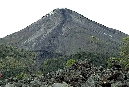 A steep volcano with a dark, rocky surface and white patches at the peak, surrounded by greenery and boulders.